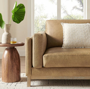 Amadeo Toast Vegan Leather Modern Sofa Beige sofa with a textured pillow in a living room setting with a wooden side table and plant.