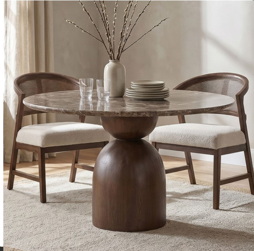 Artemos Contemporary Dark Brown Round Dining Table A round dining table with a polished stone top and a substantial dark wood pedestal base, accompanied by two matching wooden chairs with woven backs and cream upholstered seats. The table is styled with a ceramic vase of branches, glasses, and stacked plates in a softly lit dining room.