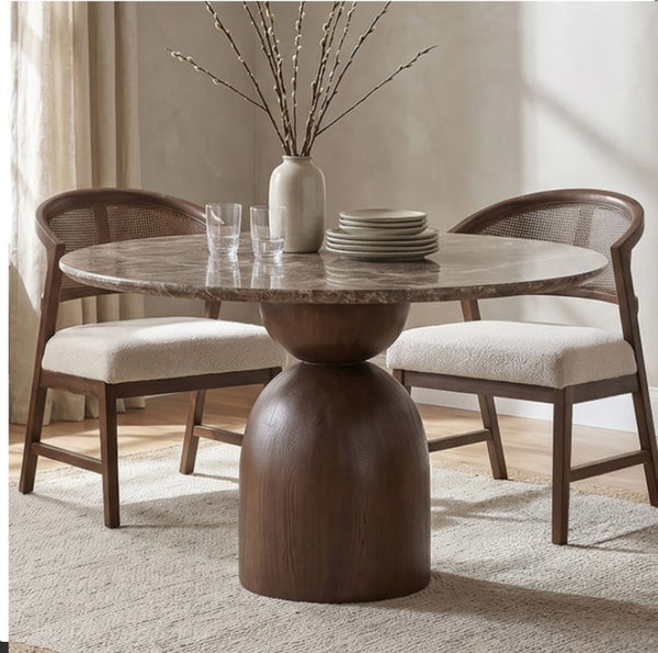 Artemos Contemporary Dark Brown Round Dining Table A round dining table with a polished stone top and a substantial dark wood pedestal base, accompanied by two matching wooden chairs with woven backs and cream upholstered seats. The table is styled with a ceramic vase of branches, glasses, and stacked plates in a softly lit dining room.