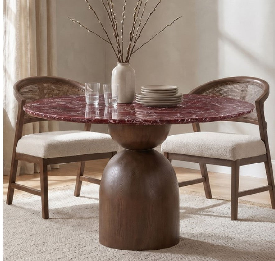 Artemos Contemporary Red Round Dining Table A round dining table with a dark reddish-brown marble top and a substantial dark wood pedestal base, surrounded by two woven-back chairs with cream upholstered seats in a softly lit room.