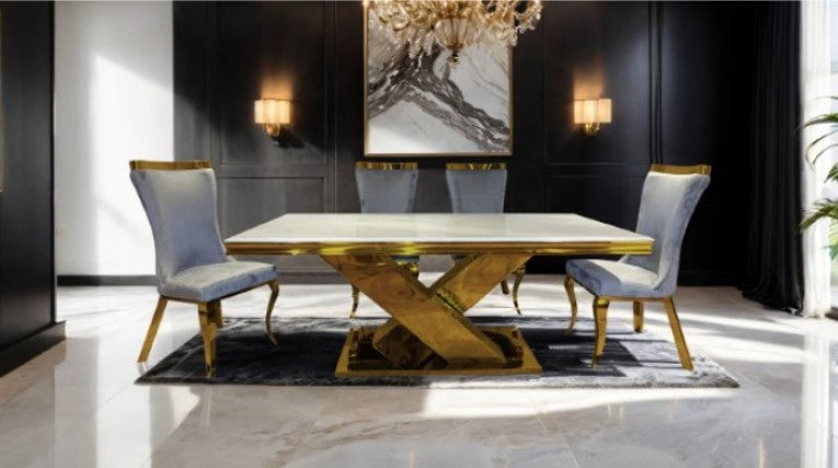 Asher Gold White Cross Shaped Glam Dining Table A luxury dining set featuring a rectangular table with a white marble top and a gold-finished geometric pedestal base, surrounded by four black upholstered chairs with gold trim, set on a round white rug in a modern room with a sea view.