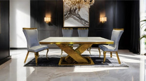 Asher Gold White Cross Shaped Glam Dining Table A luxury dining room set featuring a rectangular white marble table with a gold metallic X-shaped base, surrounded by four matching high-back chairs with light blue-gray velvet upholstery and gold trim, set on a dark patterned rug in a modern room with black paneled walls.
