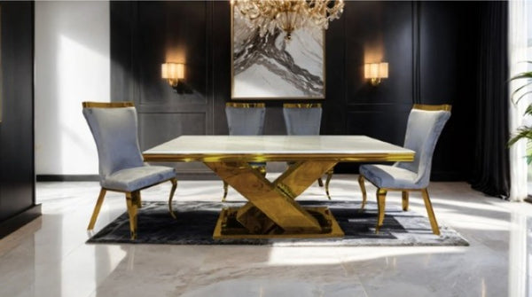 Asher Gold White Cross Shaped Glam Dining Table A luxury dining room set featuring a rectangular white marble table with a gold metallic X-shaped base, surrounded by four matching high-back chairs with light blue-gray velvet upholstery and gold trim, set on a dark patterned rug in a modern room with black paneled walls.