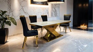 Asher Gold White Cross Shaped Glam Dining Table A modern dining room setup featuring a rectangular table with a white marble top and a reflective gold geometric base, surrounded by four black upholstered chairs with gold legs.