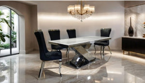 Asher Silver White Cross Shaped Glam Dining Table A modern dining room setup featuring a rectangular white marble-top dining table with a polished chrome base, surrounded by six black upholstered dining chairs with chrome legs, under a crystal chandelier.