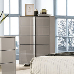 Asiko Modern UV Finish Light Taupe Chest A tall, modern 5-drawer dresser in a glossy taupe finish stands in a bright bedroom next to a bed and another matching dresser. The room features large windows with a view of bare trees outside.