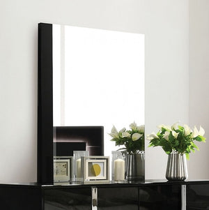 Asiko Modern UV Finish Piano Black Mirror A large rectangular wall mirror with a slim black frame hangs on a white wall above a glossy black console table. The mirror reflects a dark object, while the room setting includes two vases with white flowers and small framed decor on the table.