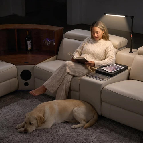 Aurelia Beige Modern Air Leather Sectional Sofa Powered Recliner A woman relaxes in a cream leather sectional sofa while reading, with a yellow Labrador retriever lying on the gray carpet on the floor. The scene is set in a dimly lit living room with a wooden side table and a floor lamp.
