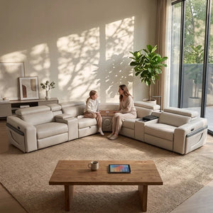 Aurelia Beige Modern Air Leather Sectional Sofa Powered Recliner A modern U-shaped sectional sofa in light beige leather is arranged in a bright living room with a woman and child sitting in the center, facing a wooden coffee table. The room features warm natural light casting leaf shadows on the wall, a large potted plant, and a large window in the background.