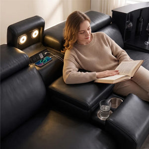Aurelia Modern Air Leather Sectional Sofa Powered Recliner A woman relaxes in a black leather sectional sofa, reading a book. The sofa features a headrest with built-in lights, a charging port with a phone plugged in, and a cup holder with a glass of water.