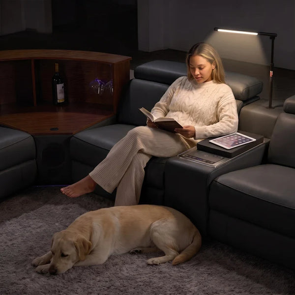 Aurelia Modern Air Leather Sectional Sofa Powered Recliner A woman relaxes in a dark leather reclining sofa while reading a book, with a yellow Labrador retriever lying on a shag rug in the foreground. A floor lamp illuminates the scene, and a wine bottle and glasses are visible in a wooden cabinet in the background.