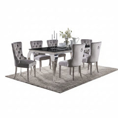 Betsy Silver Black Glass Top Modern Dining Room Table A 7-piece dining set featuring a rectangular glass-top table with a polished silver frame and six matching tufted gray velvet chairs, styled on a shag rug in a modern dining room.