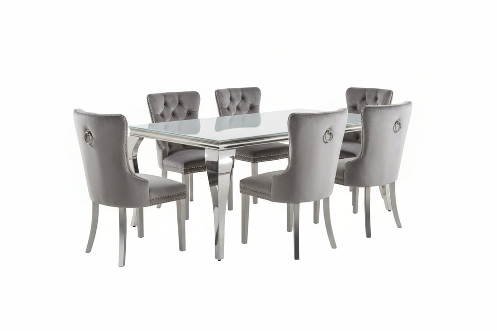 Betsy Silver White Glass Top Modern Dining Room Table A 7-piece dining room set featuring a rectangular glass-top table and six tufted gray velvet chairs with silver legs, arranged on a shag rug in a modern dining room.