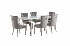 Betsy Silver White Glass Top Modern Dining Room Table A 7-piece dining room set featuring a rectangular glass-top table and six tufted gray velvet chairs with silver legs, arranged on a shag rug in a modern dining room.