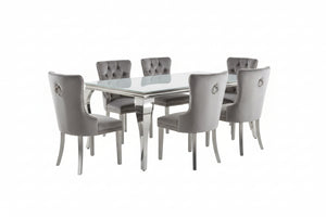 Betsy Silver White Glass Top Modern Dining Room Table A 7-piece dining room set featuring a rectangular glass-top table and six tufted gray velvet chairs with silver legs, arranged on a shag rug in a modern dining room.