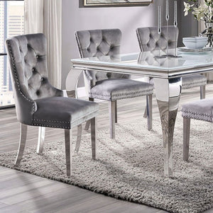Betsy Silver White Glass Top Modern Dining Room Table A modern dining set featuring a rectangular glass-top table with a polished silver metal frame and four matching tufted gray velvet chairs with silver nailhead trim, set on a shag rug in a bright room.