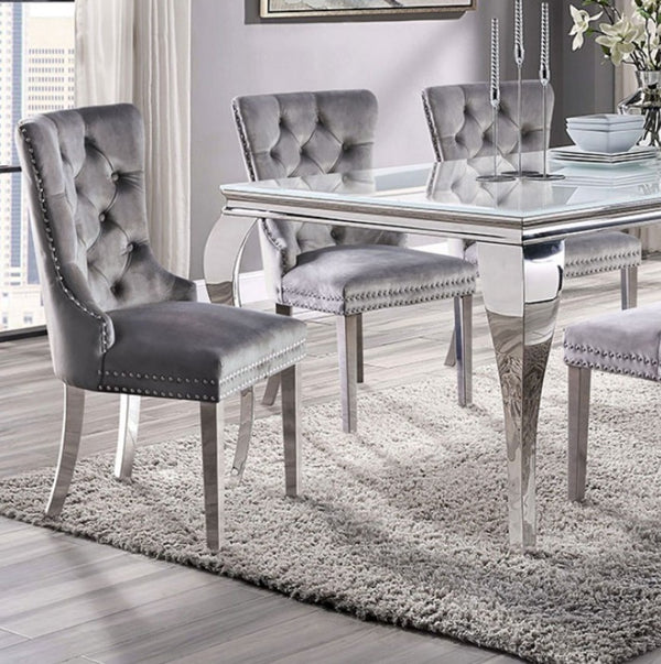 Betsy Silver White Glass Top Modern Dining Room Table A modern dining set featuring a rectangular glass-top table with a polished silver metal frame and four matching tufted gray velvet chairs with silver nailhead trim, set on a shag rug in a bright room.