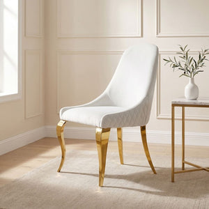 Betsy White Upholstered Modern Chair (2/CTN) A modern dining chair with a white quilted upholstered seat and backrest, supported by elegant curved gold metal legs, set in a bright, elegant room next to a marble-topped side table.