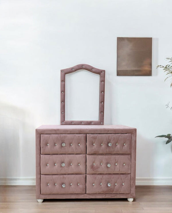 Chloe Pink Fabric Full Upholstered Kids Dresser A dusty pink velvet dresser with six drawers, tufted details, and silver crystal knobs, featuring an arched matching mirror on top. The dresser is set against a white wall with a small framed piece of art and a plant in the background.