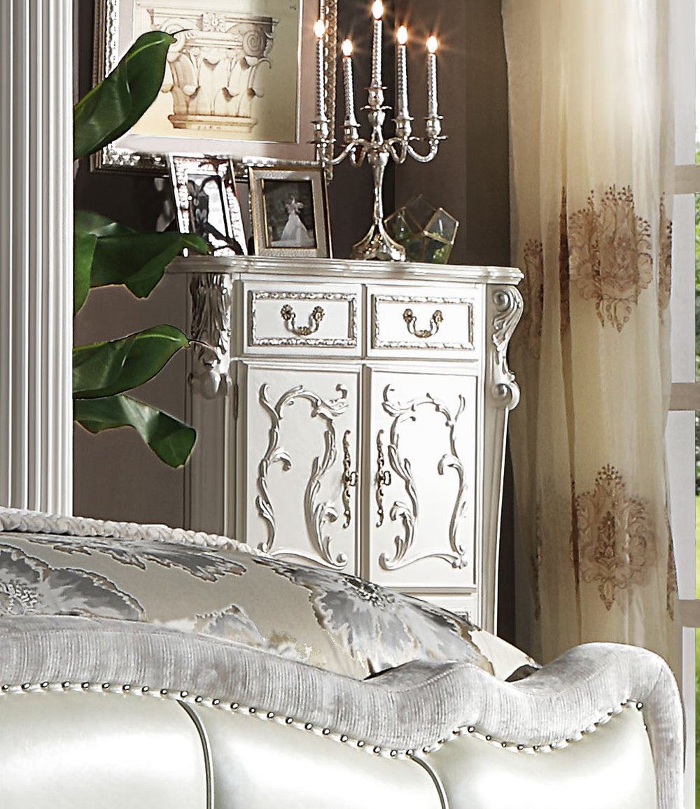 Dresden Bone White Traditional Style Chest An ornate white carved wooden cabinet or sideboard in a styled bedroom setting, with a silver candelabra on top, framed photos, a mirror, curtains, and part of a tufted upholstered bed visible in the foreground.