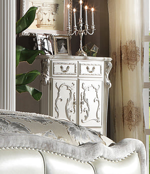 Dresden Bone White Traditional Style Chest An ornate white carved wooden cabinet or sideboard in a styled bedroom setting, with a silver candelabra on top, framed photos, a mirror, curtains, and part of a tufted upholstered bed visible in the foreground.