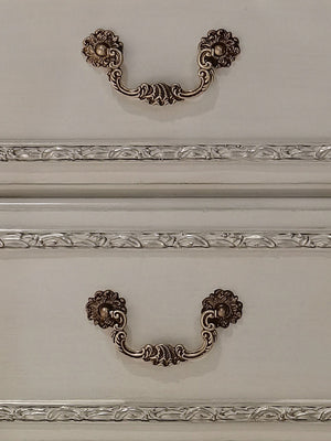 Dresden Bone White Traditional Style Nightstand Two ornate antique bronze drawer pulls with floral scrollwork and decorative medallions, mounted on a cream-colored wooden cabinet with decorative molding.