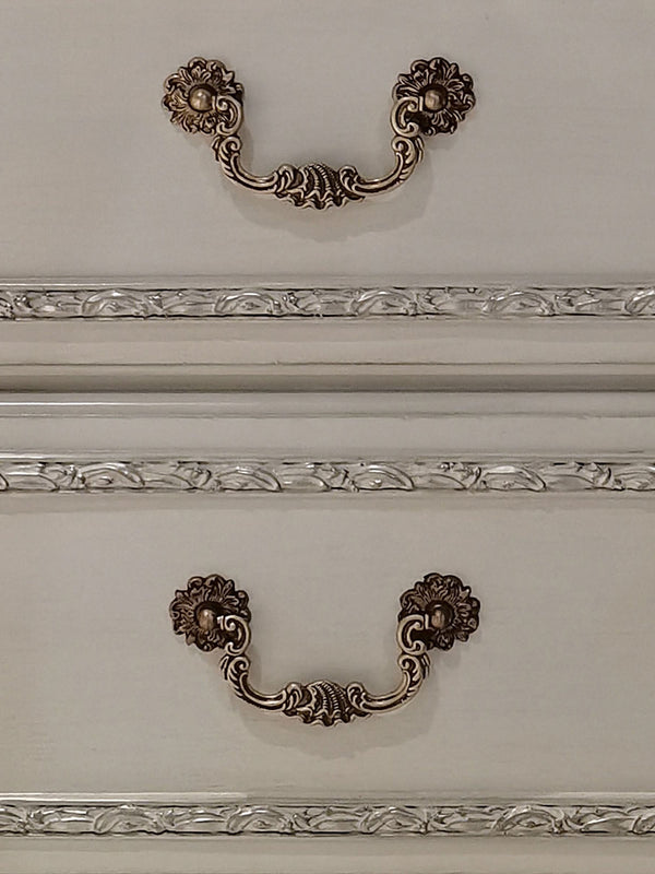 Dresden Bone White Traditional Style Nightstand Two ornate antique bronze drawer pulls with floral scrollwork and decorative medallions, mounted on a cream-colored wooden cabinet with decorative molding.