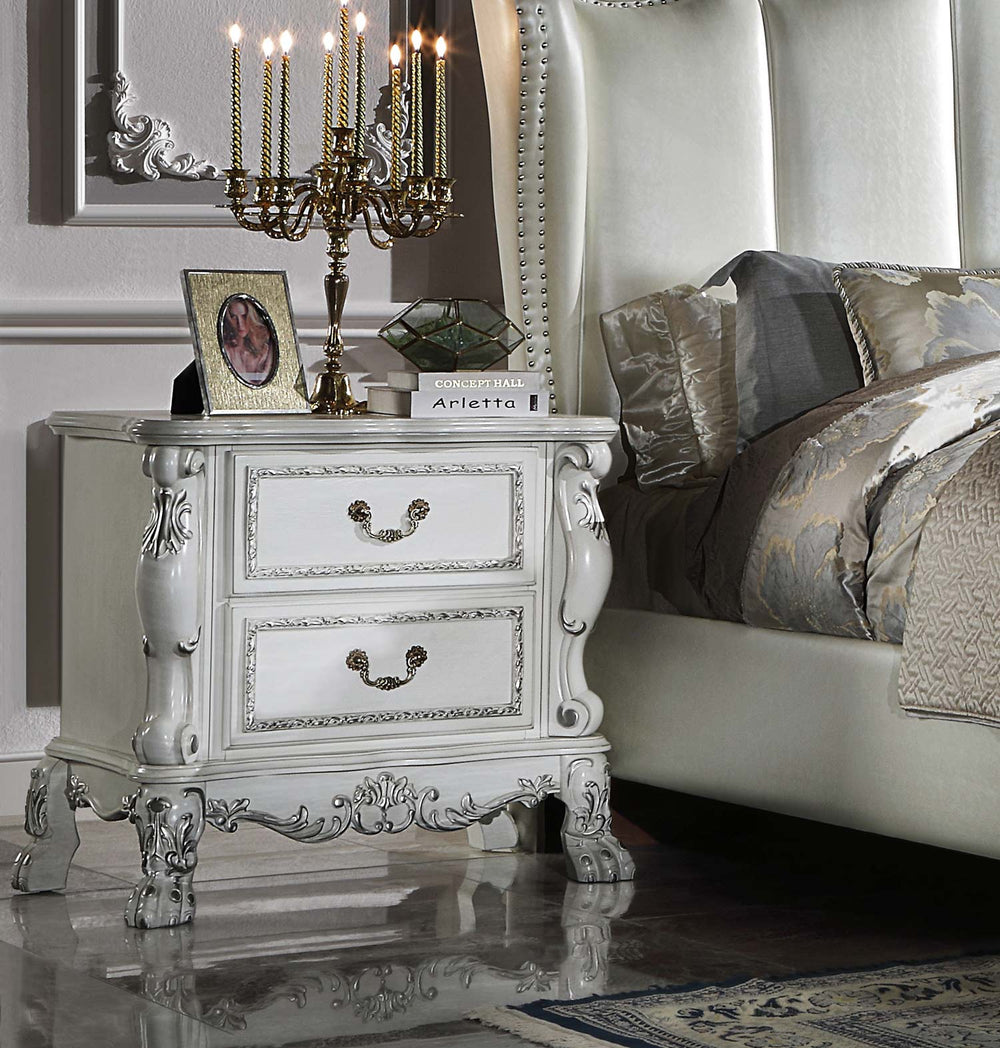 Dresden Bone White Traditional Style Nightstand An ornate white two-drawer nightstand with carved details and dark metal handles, styled in a bedroom next to a bed with gray and beige patterned bedding. The nightstand is decorated with a gold candelabra, a picture frame, and a small book.