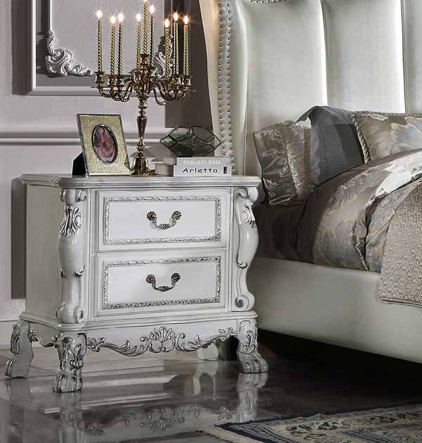 Dresden Bone White Traditional Style Nightstand An ornate white two-drawer nightstand with carved details and dark metal handles, styled in a bedroom next to a bed with gray and beige patterned bedding. The nightstand is decorated with a gold candelabra, a picture frame, and a small book.