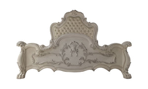 Dresden Bone White Traditional Silhouette Upholstered Bed An ornate, vintage-style upholstered headboard in a distressed off-white or cream color, featuring intricate carved floral details and a tufted button pattern on the top panel.
