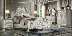 Dresden Bone White Traditional Silhouette Leaf Carving Inlay Bed An ornate white carved bedroom furniture set in a luxury bedroom, featuring a bed with a high sculpted headboard and footboard, a matching nightstand, dresser, and mirror, all styled with gray and beige bedding and a blue patterned rug.