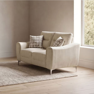 Estrella Light Taupe Brown Slope Flair Arm Loveseat A light beige upholstered loveseat with two patterned throw pillows, placed on a woven rug in a bright room with wood flooring and a window.