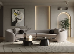 Kilmer Gray Fabric Curved Modern Sectional Sofa A modern curved light gray sofa in a minimalist living room setting, styled with black accent pillows, a patterned area rug, low black coffee tables, and a framed abstract art print on the wall.