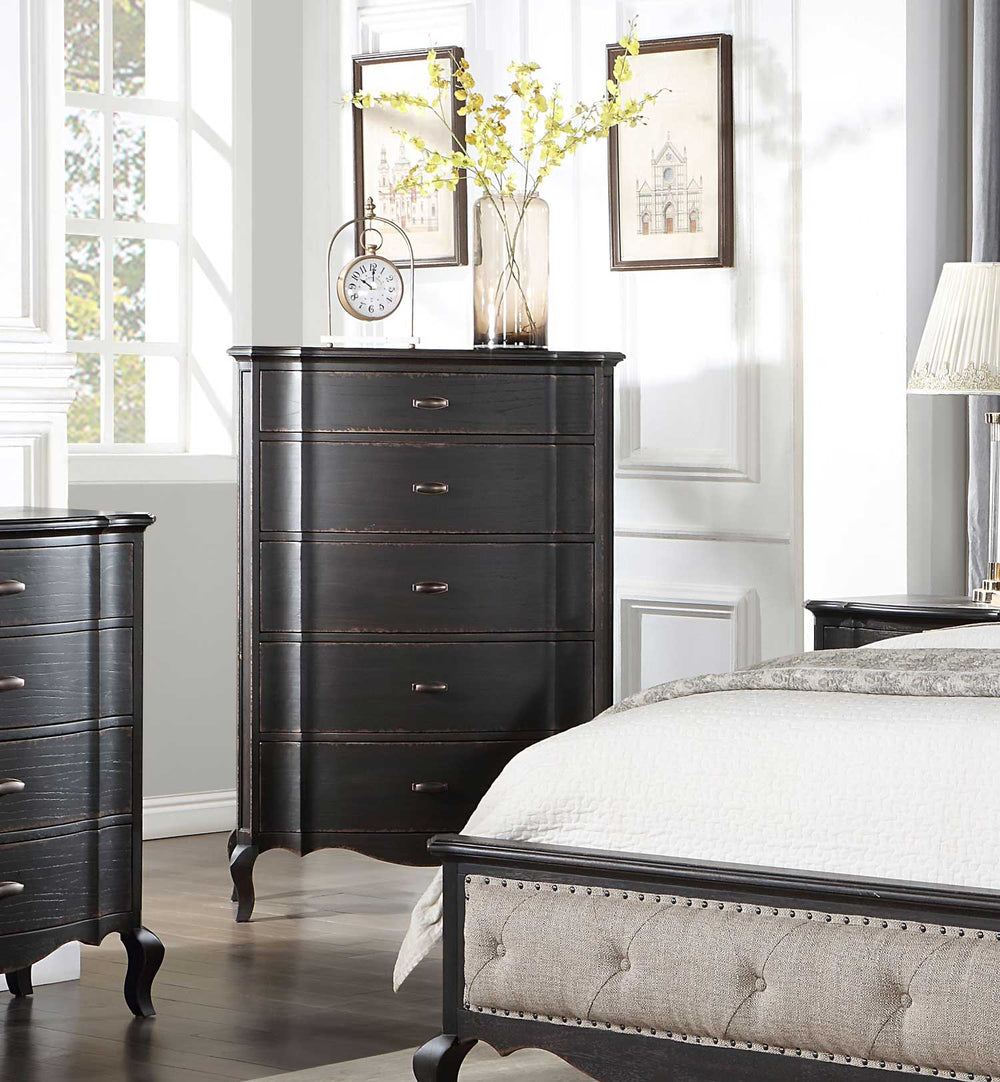 Lesley Dark Brown Chest A dark wood five-drawer chest of drawers in a bedroom setting, styled with a vase of yellow flowers, a small clock, and framed prints on top, next to a bed and dresser.