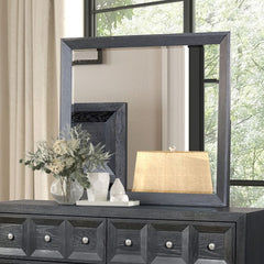 Montague Cross Modern Classic Weathered Black Mirror A large rectangular wall mirror with a dark charcoal gray rustic wood frame, styled on top of a matching dark gray dresser in a bedroom setting. The dresser features decorative drawer fronts with silver knobs, a beige lamp with a linen shade, and a vase of dried flowers.