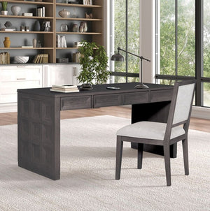 Montague Cross Modern Style Black Writing Desk A dark gray wooden writing desk with three drawers and a paneled front leg, paired with a matching gray upholstered chair, set in a bright home office with a bookshelf and large windows.