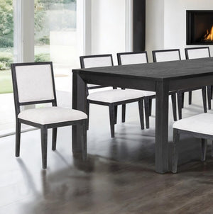 Montague Cross Modern Style Weathered Black Rectangular Dining Table A dark wood dining table with six matching chairs featuring dark frames and light beige upholstered seats, set in a modern dining room with a fireplace and large windows.
