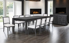 Montague Cross Modern Style Weathered Black Rectangular Dining Table A modern dining room set featuring a rectangular dark wood table and eight matching chairs with white upholstered seats, shown in a contemporary room with a fireplace and dark wood floors.