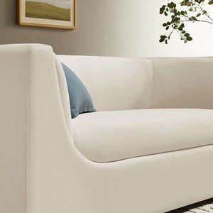 Natalia Alabaster Velvet Fabric Loveseat Beige sofa with a blue pillow in a room with a painting and plant on the wall.