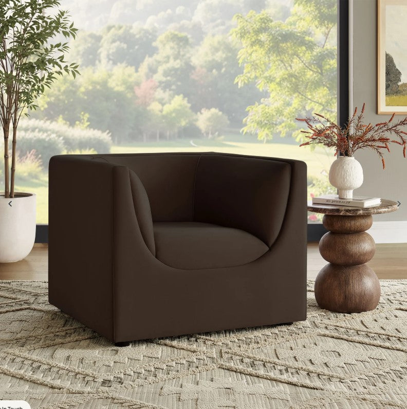 Natalia Chocolate Brown Velvet Fabric Chair Brown armchair in a room with a window view of greenery, a side table, and decorative elements.