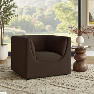 Natalia Chocolate Brown Velvet Fabric Chair Brown armchair in a room with a window view of greenery, a side table, and decorative elements.
