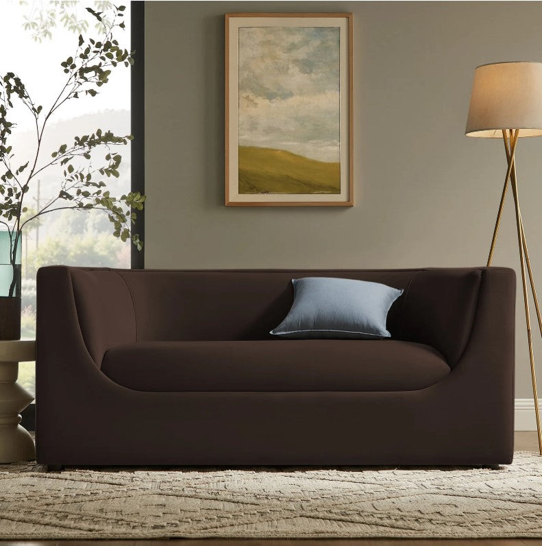 Natalia Chocolate Brown Velvet Fabric Loveseat Brown sofa with a blue pillow in a living room setting with a painting and lamp.