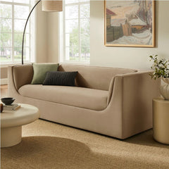 Natalia Taupe Velvet Fabric Sofa Beige sofa in a living room with a painting on the wall and a plant on a side table.