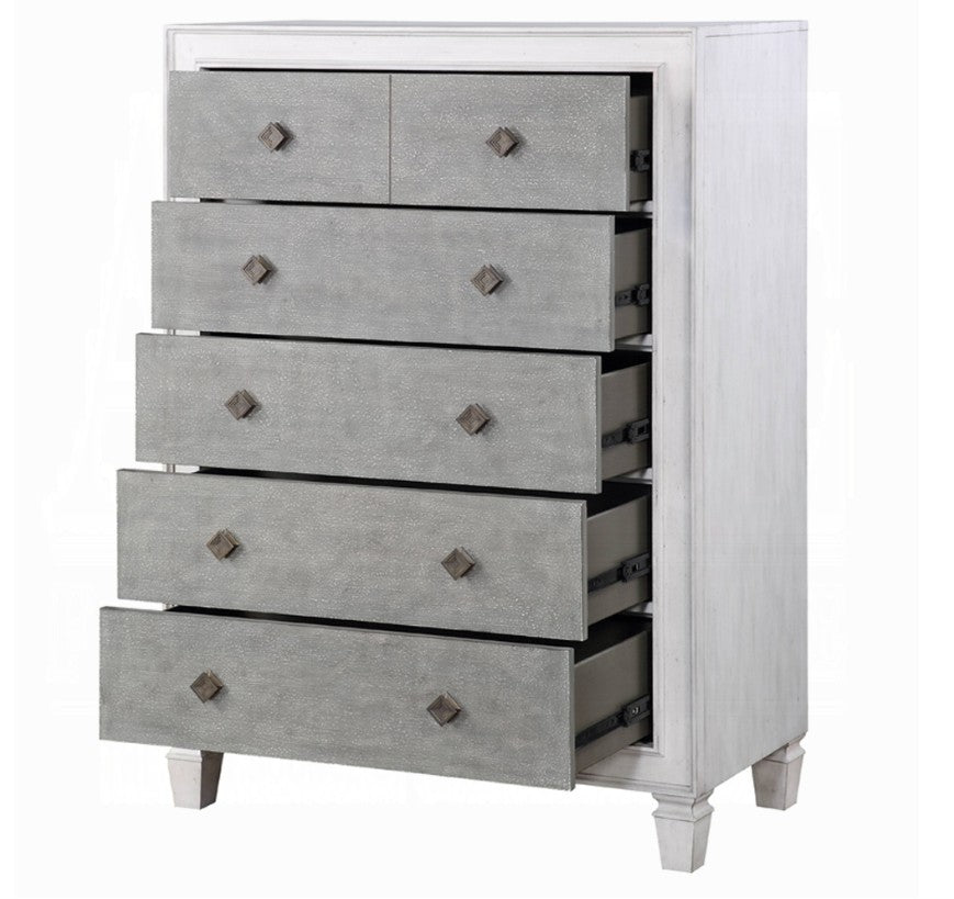 Natasha Two Tone Light Gray Chest A white five-drawer chest of drawers with textured gray front panels stands in a bedroom setting against a light gray wall, with a bed and white curtain partially visible on the right.
