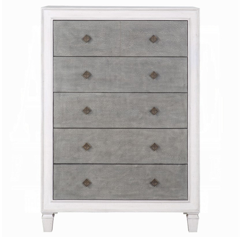 Natasha Two Tone Light Gray Chest A white five-drawer chest of drawers with textured gray front panels stands in a bedroom setting against a light gray wall, with a bed and white curtain partially visible on the right.