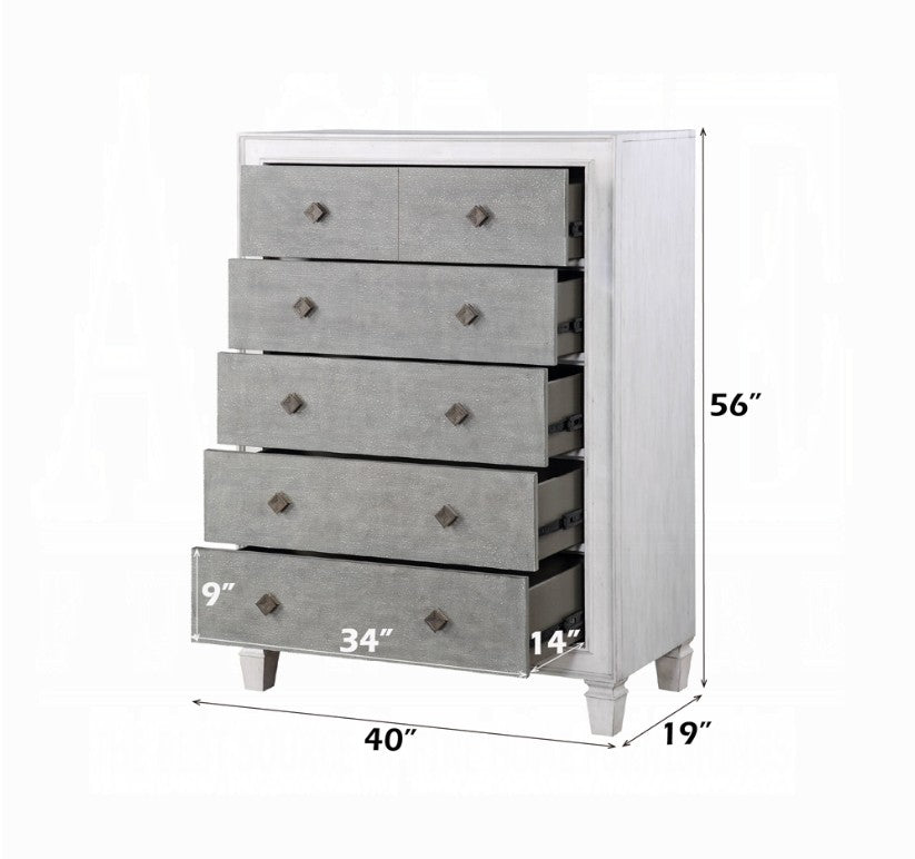 Natasha Two Tone Light Gray Chest A white five-drawer chest of drawers with textured gray front panels stands in a bedroom setting against a light gray wall, with a bed and white curtain partially visible on the right.