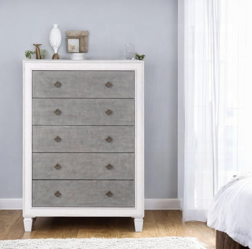 Natasha Two Tone Light Gray Chest A white five-drawer chest of drawers with textured gray front panels stands in a bedroom setting against a light gray wall, with a bed and white curtain partially visible on the right.
