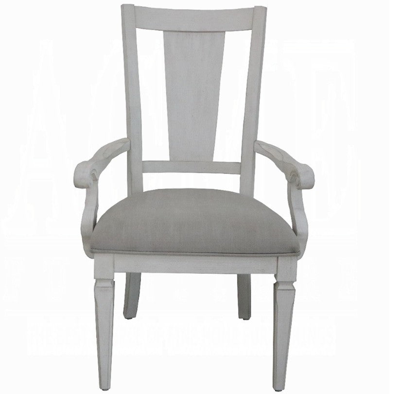 A white wooden dining armchair with a light beige upholstered seat cushion, positioned on a blue and white patterned rug in a bright room with gray curtains.