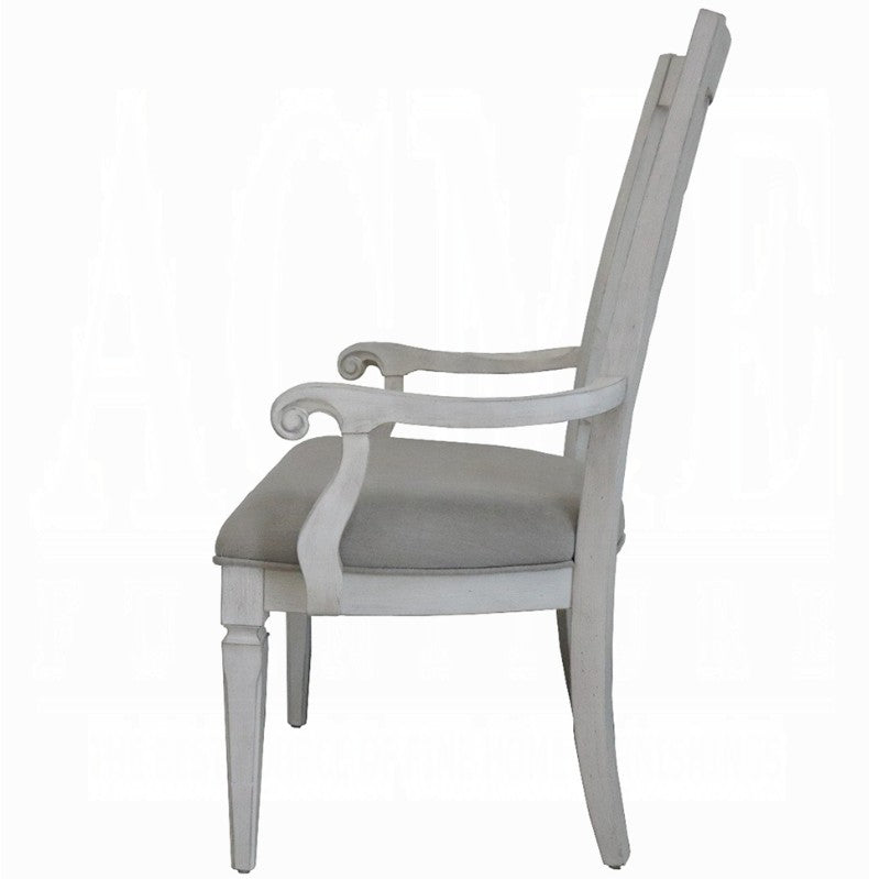 A white wooden dining armchair with a light beige upholstered seat cushion, positioned on a blue and white patterned rug in a bright room with gray curtains.