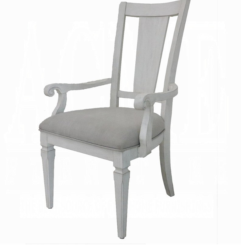 A white wooden dining armchair with a light beige upholstered seat cushion, positioned on a blue and white patterned rug in a bright room with gray curtains.